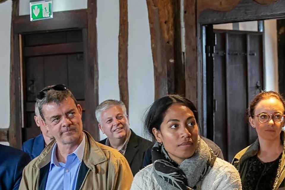 A group of people seated indoors in a room with wooden beams, listening attentively to someone off-frame.