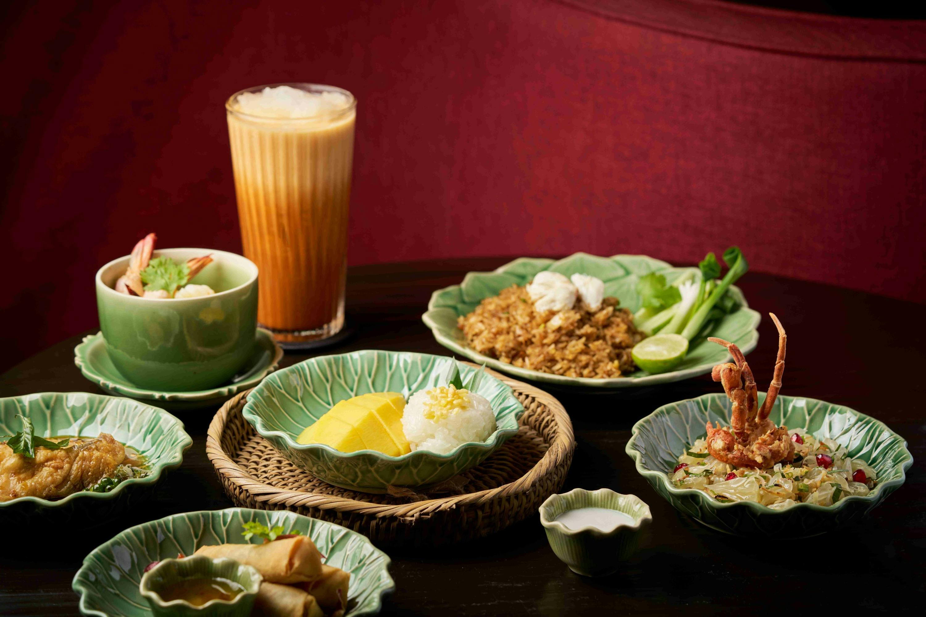 A table with various dishes including mango sticky rice, fried rice, spring rolls, and a tall glass of iced tea.