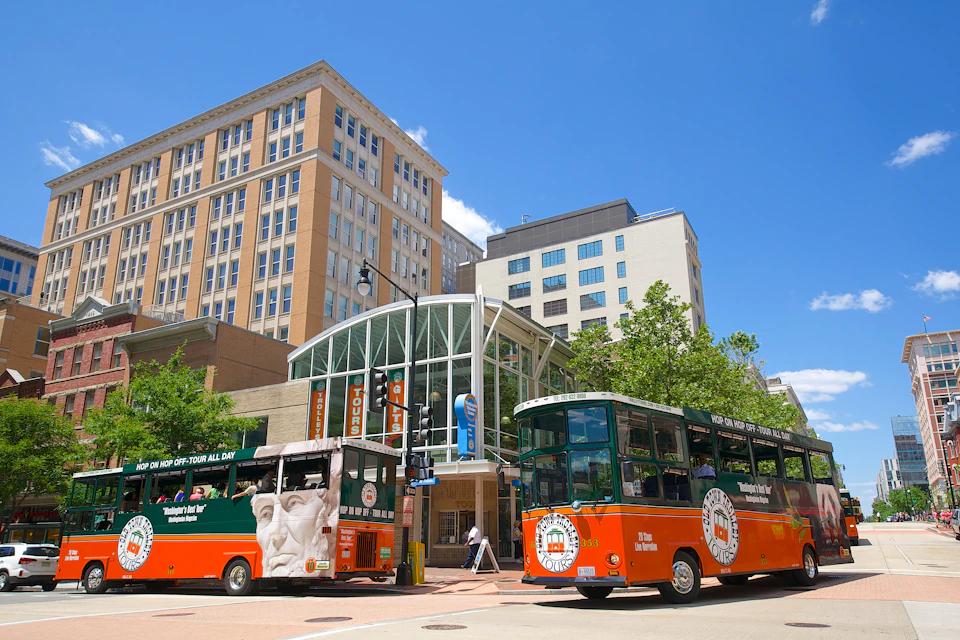 Two green and orange tour buses near a modern glass building and tall offices on a clear, sunny day.