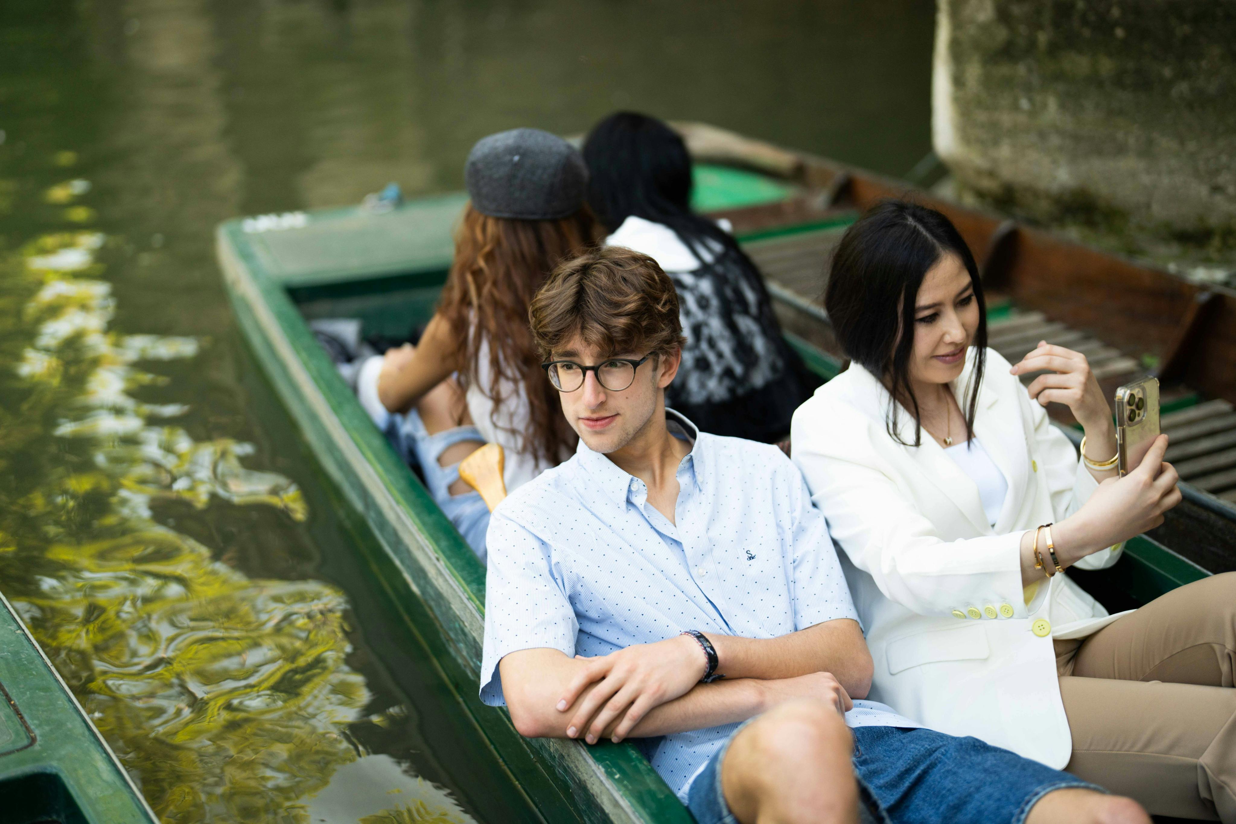Zwei Personen sitzen in einem grünen Boot, ein Mann in einem blauen Hemd und eine Frau in einer weißen Jacke, die ein Selfie machen. Dahinter sitzen zwei weitere Personen.