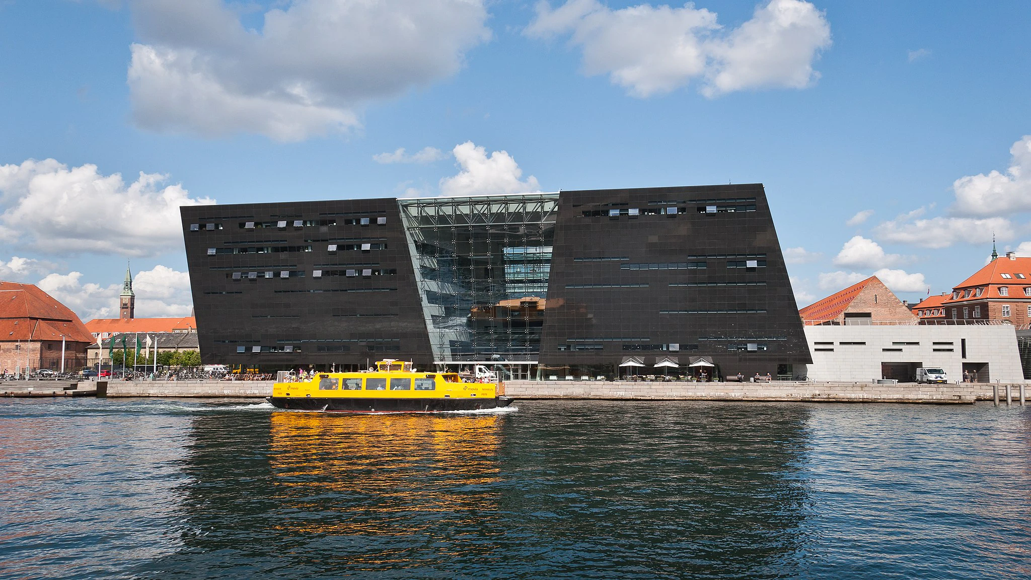 Modern svart byggnad med sneda väggar, intilliggande äldre konstruktioner med röda tak, gul färja på vattnet i förgrunden.