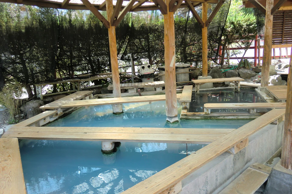 An outdoor hot spring pool with wooden walkways and a shade structure, surrounded by vegetation and privacy screens.