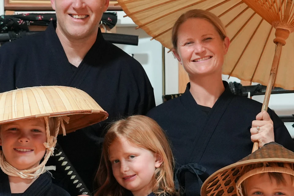 A family dressed in traditional Japanese clothing, holding swords and umbrellas, standing in front of a display of swords.