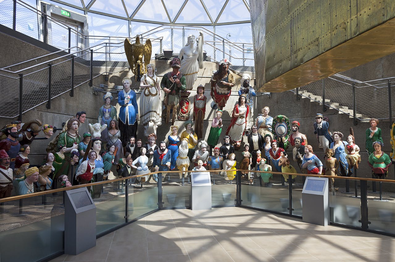 A display of colorful maritime figureheads in a museum setting, with a glass ceiling and informational plaques in the foreground.