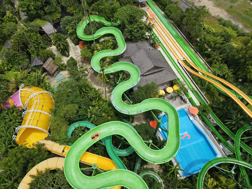 Aerial view of a water park with winding green slides, a pool, and surrounding greenery and structures.