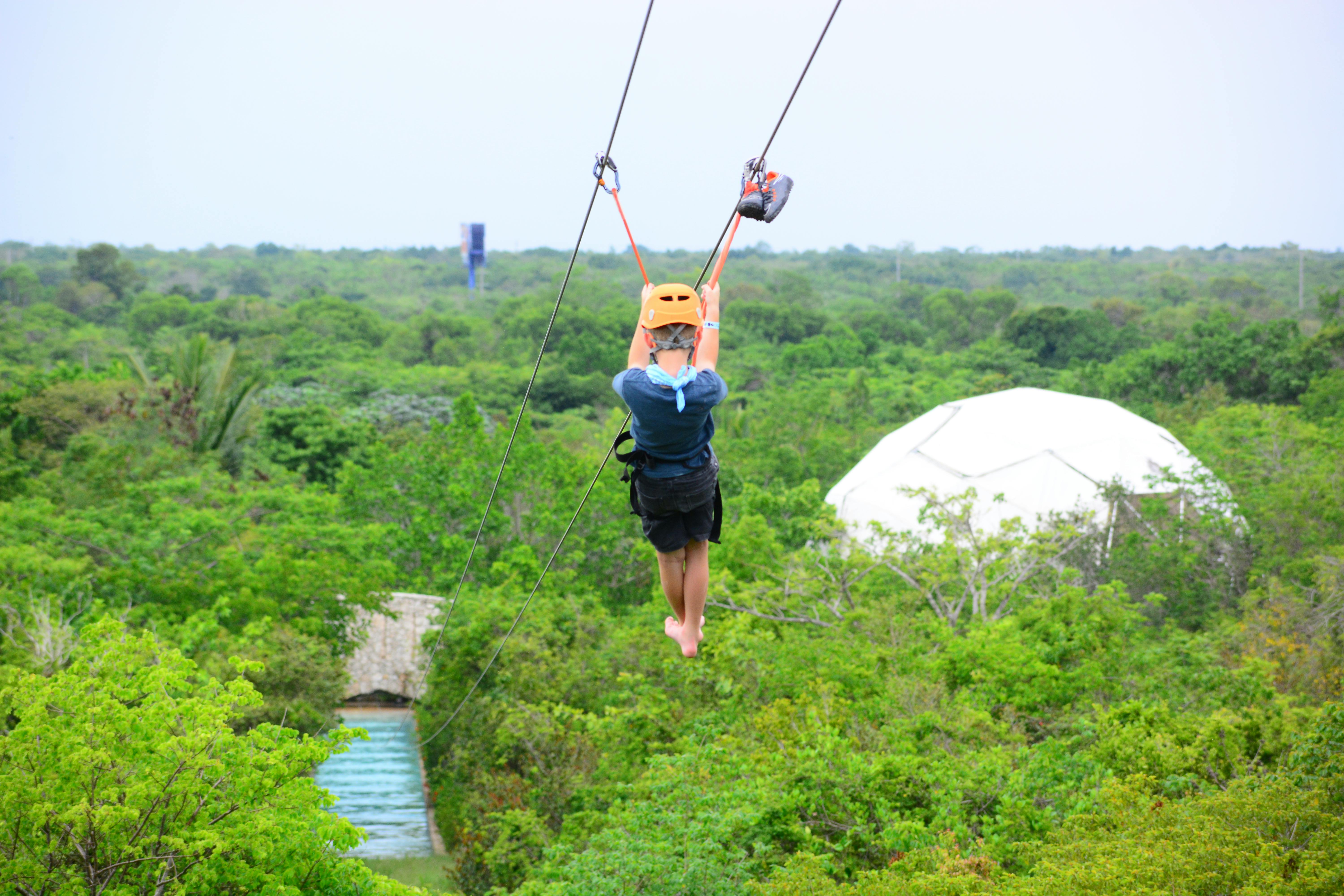 青いシャツ、黒いショーツ、黄色いヘルメットを着て、緑豊かな森の上にジップライニングをしている人。 背景に白いドーム構造。