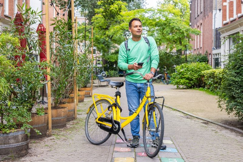 A man in a green sweater holds a phone and stands next to a yellow bicycle on a path in a green, urban park.