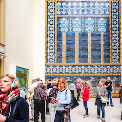 Människor som utforskar ett museum med stora, invecklade blå och gula mosaikväggar och olika andra konstverk som visas. Många bär ljudguider.