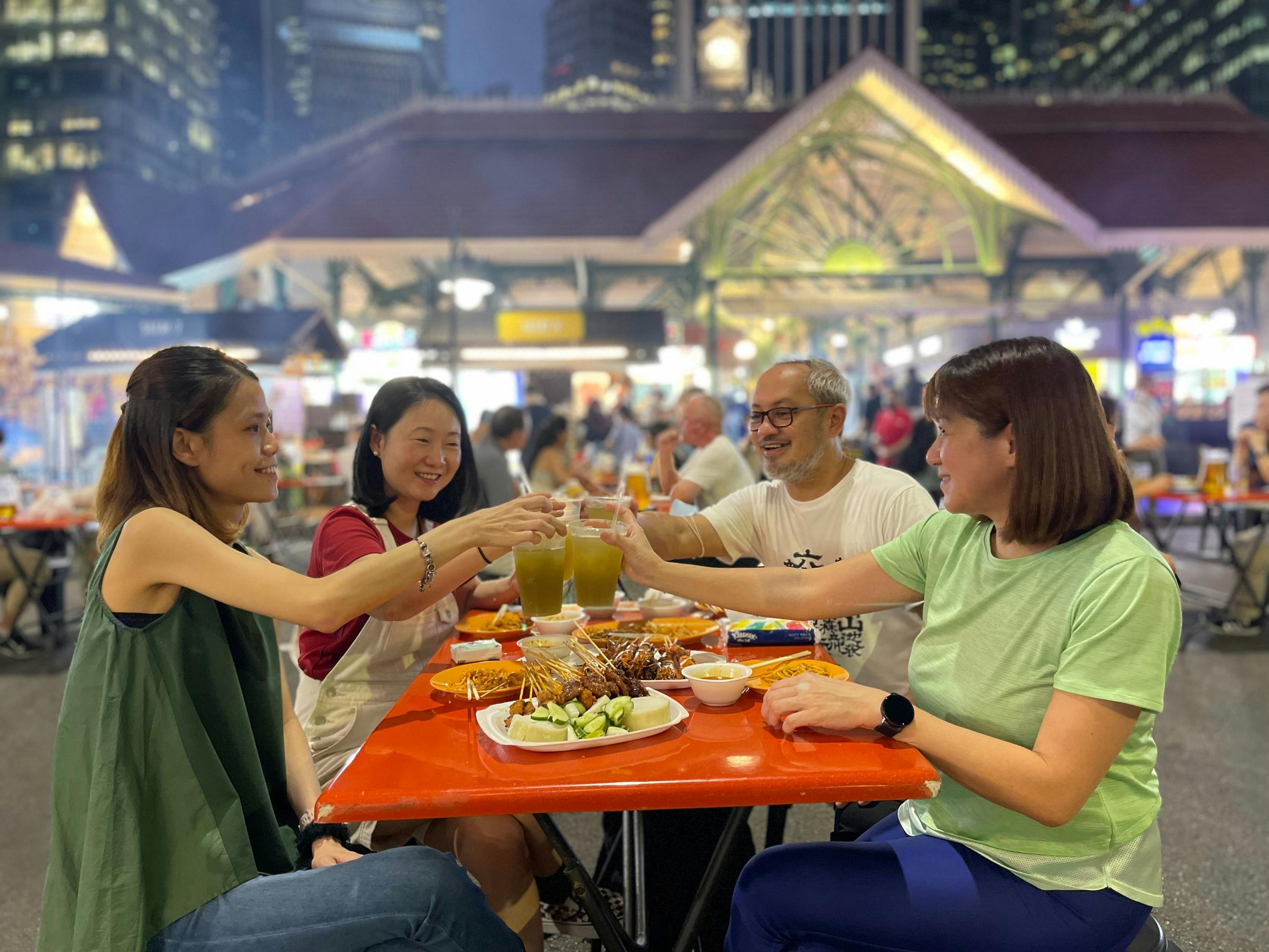 A group of people sitting at an outdoor table in a food market, clinking glasses over a meal, with urban buildings in the background.