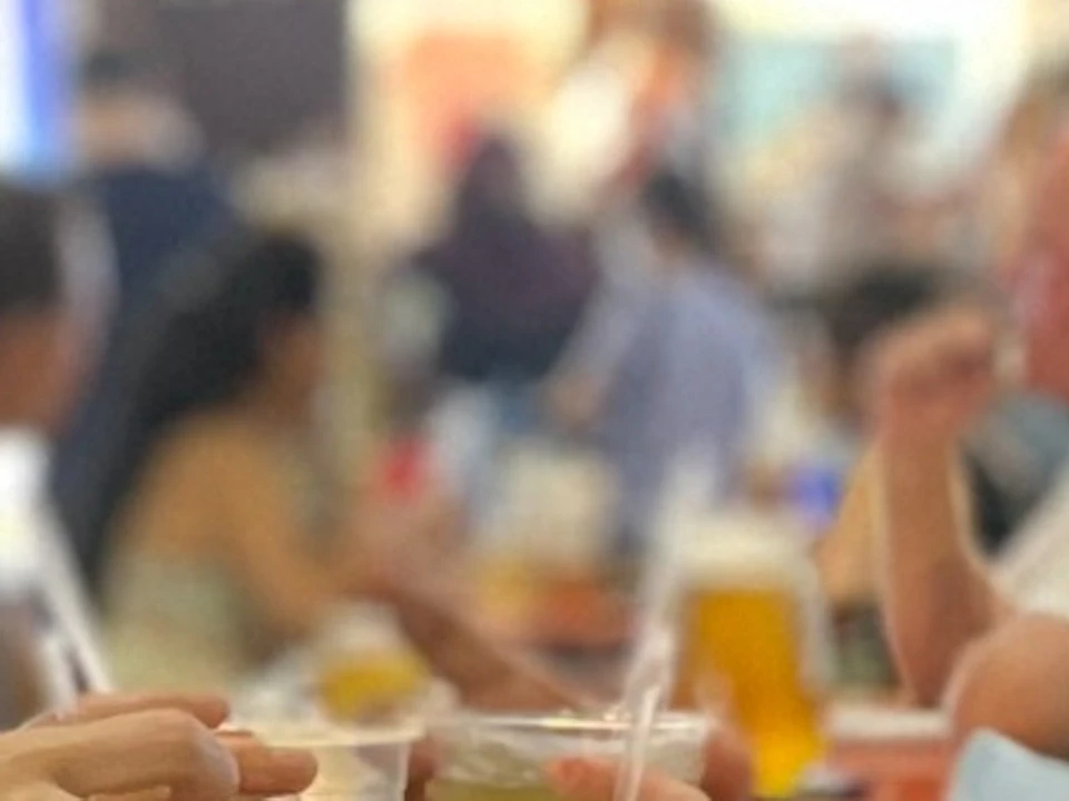 A group of people sitting at an outdoor table in a food market, clinking glasses over a meal, with urban buildings in the background.