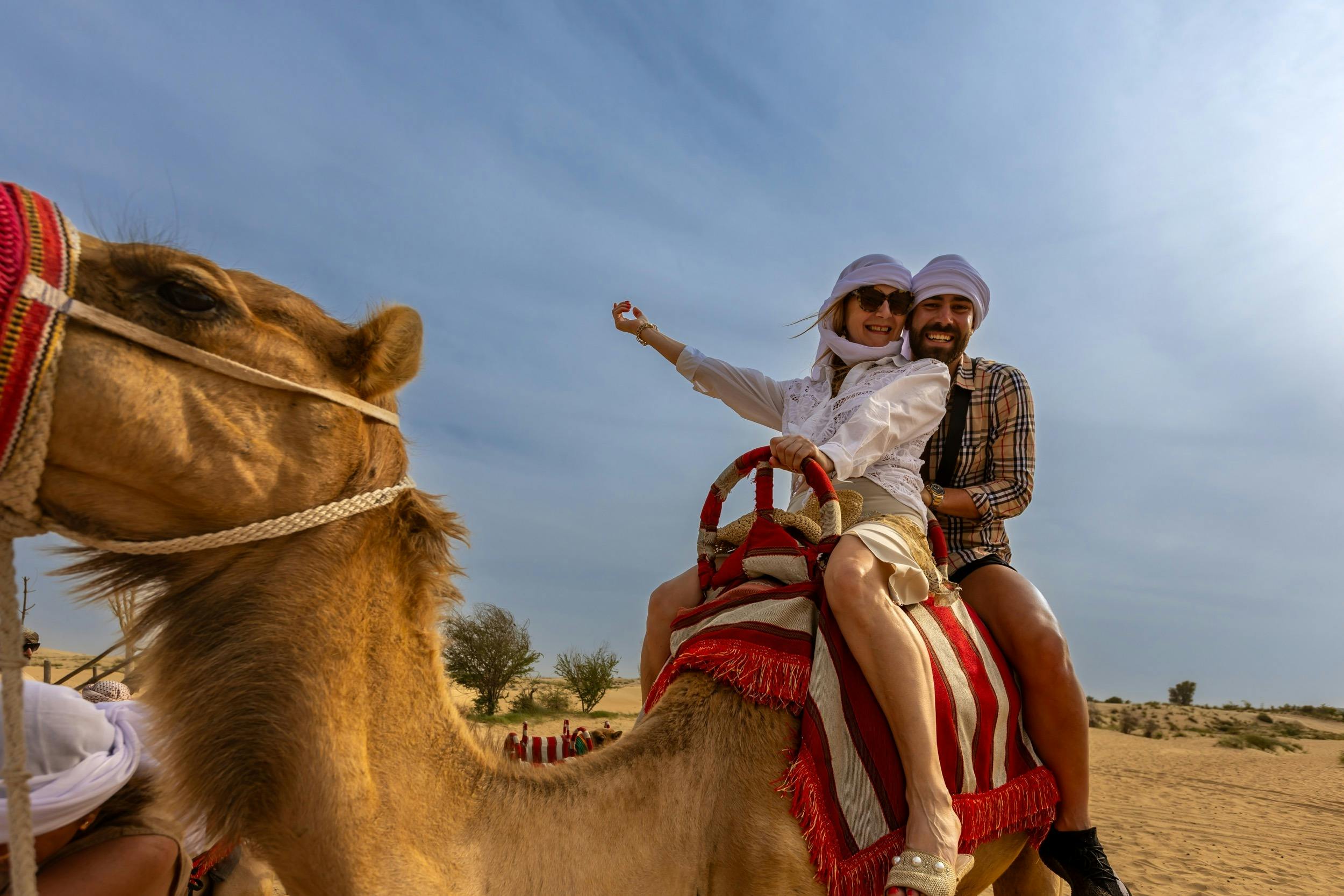 Two people wearing headscarves ride a camel with a red and white saddle in a desert landscape under a blue sky.
