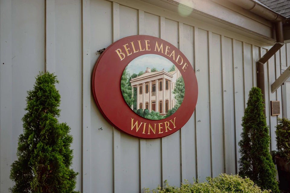 A round, red sign with "Belle Meade Winery" in gold lettering and an illustration of a building, mounted on a gray, wood-paneled wall.