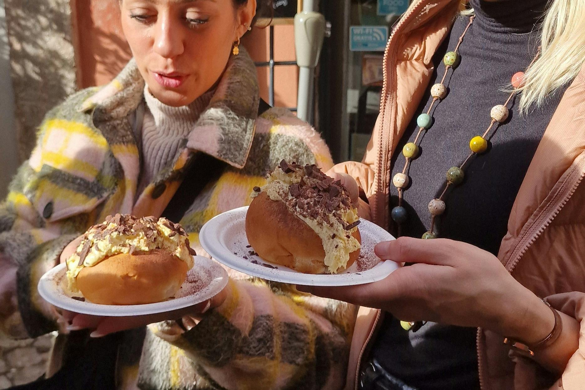 A woman in a checkered coat holds two plates with cream-filled pastries topped with chocolate shavings.