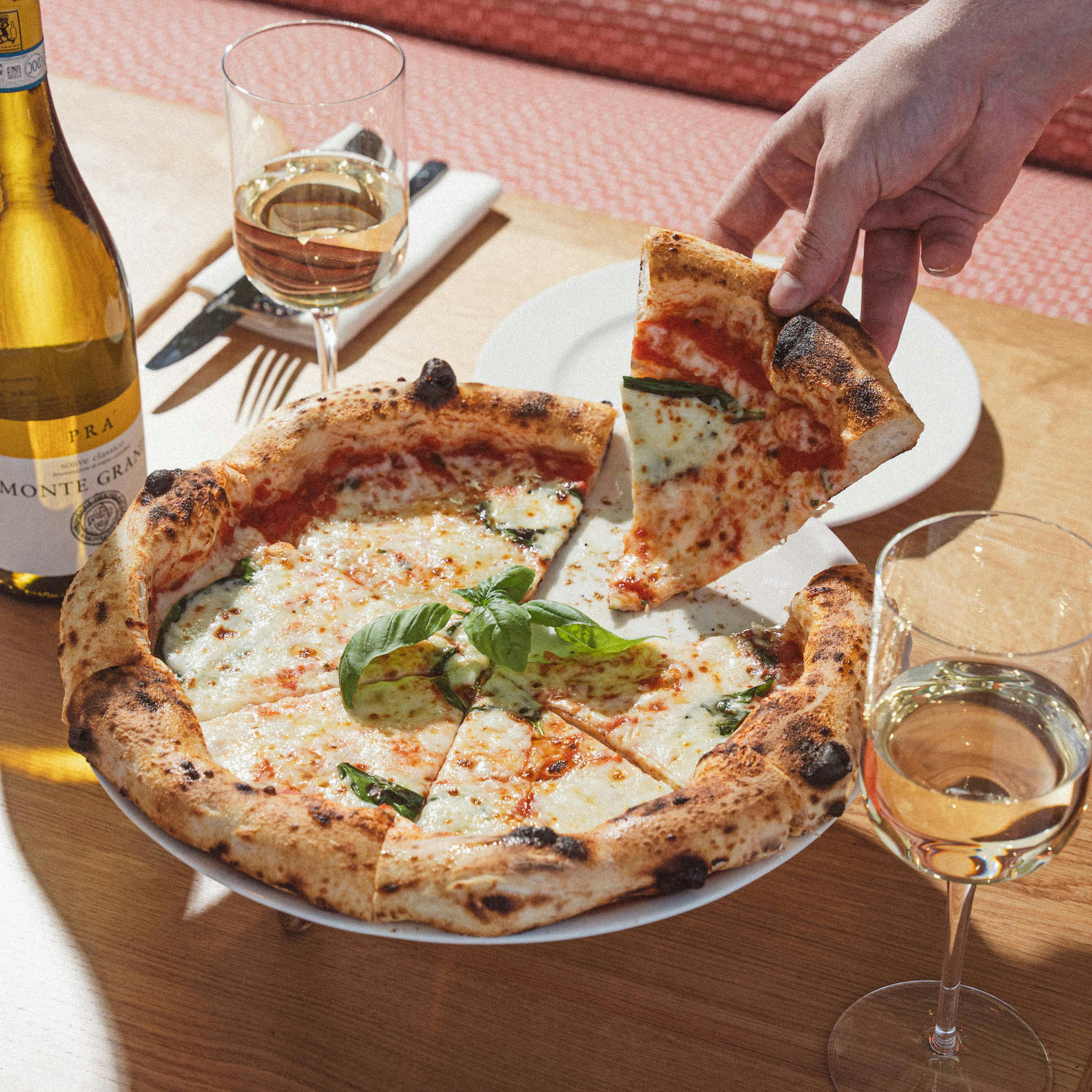 Una mano solleva un trancio di pizza Margherita da un tavolo di legno apparecchiato con una bottiglia di vino, due bicchieri di vino bianco e degli utensili.