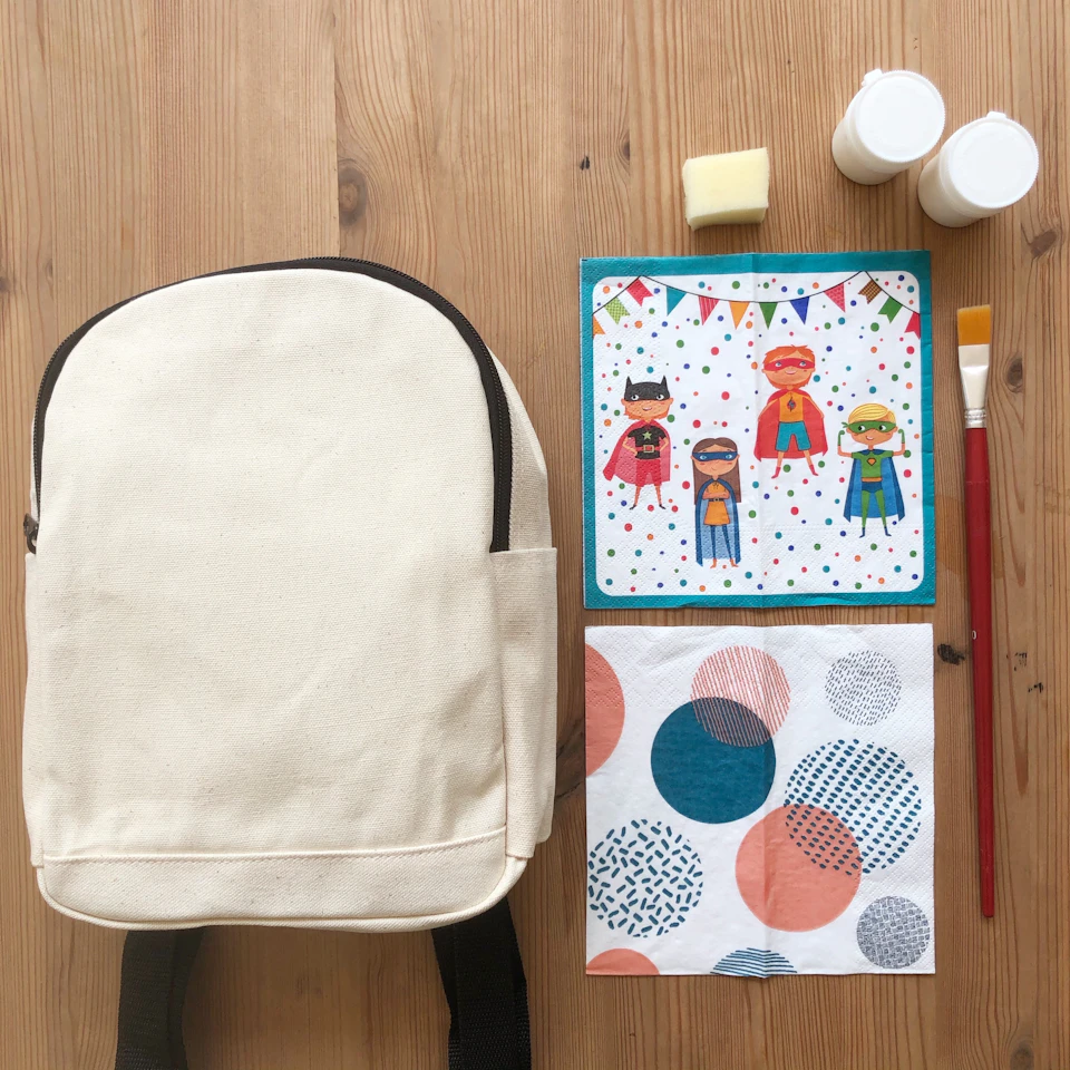A crafting kit featuring a blank canvas backpack, paint supplies, and colorful decorative materials on a wooden surface.