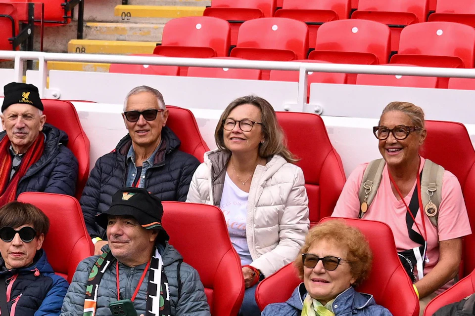 A group of people sitting on red stadium seats near a sports field, listening to someone speaking to them.