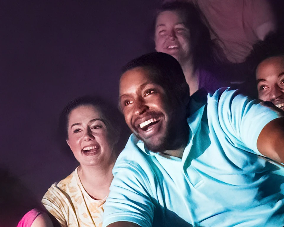 People smiling and laughing as they ride a roller coaster in a dimly lit environment.