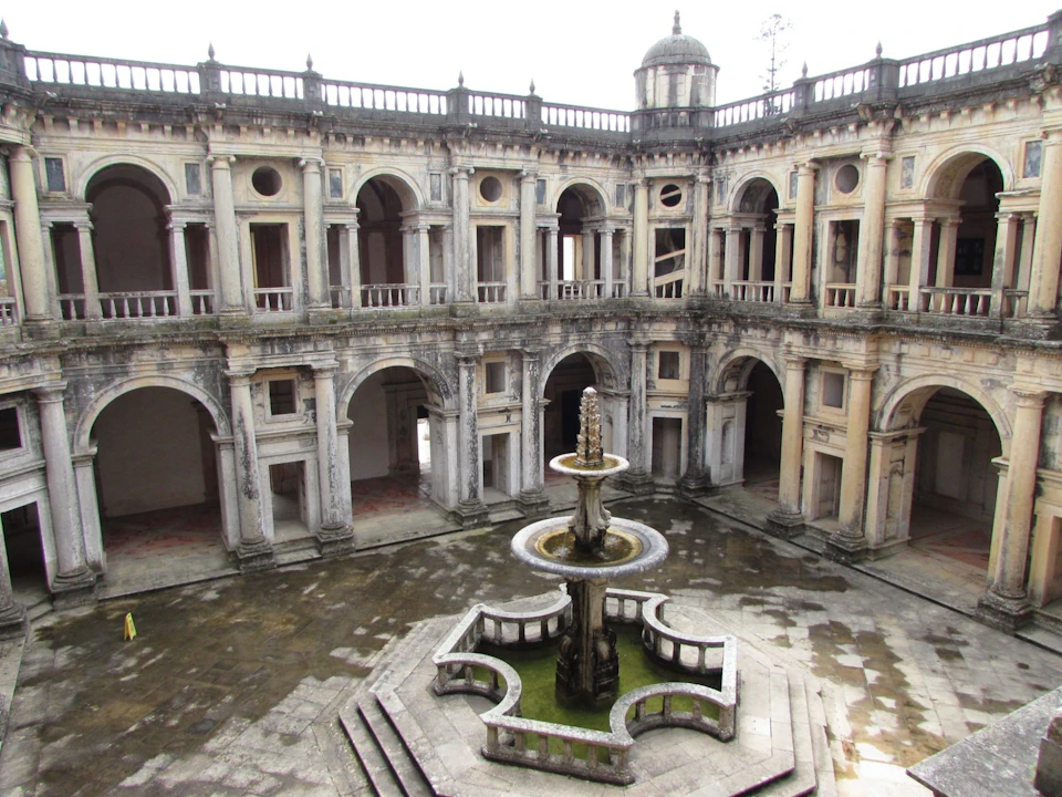 An old stone courtyard featuring an ornate tiered fountain at the center, surrounded by a two-story building with arched windows and doorways.