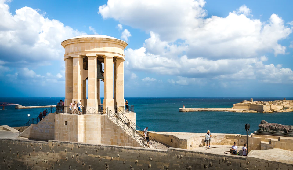 People walking around a stone rotunda structure overlooking a blue sea and cloudy sky.