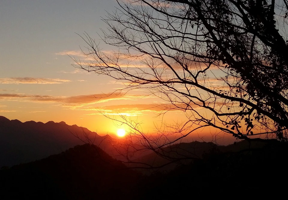 A scenic sunrise over a mountain range with a tree in the foreground and vibrant orange and yellow hues in the sky.