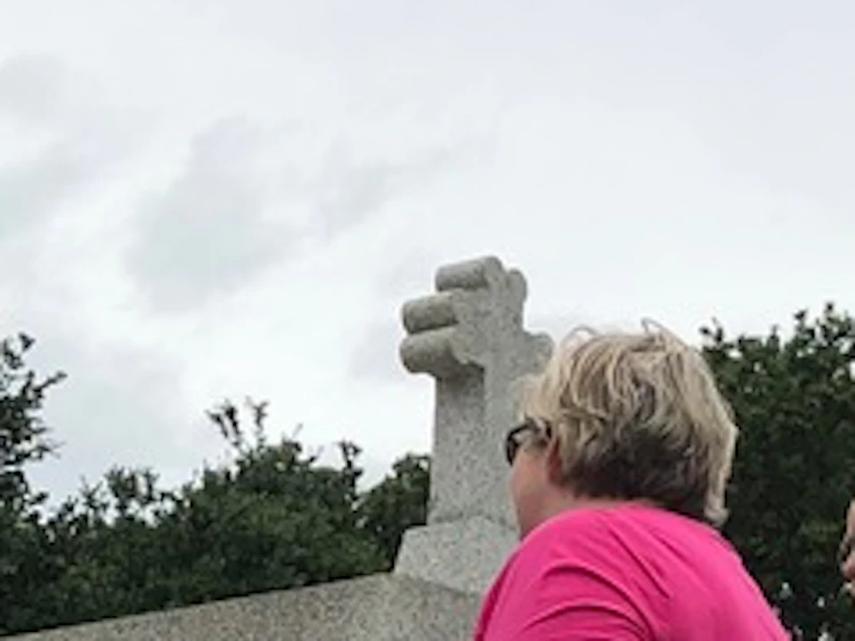 A group of people is gathered in front of a white mausoleum with a cross on top, listening to a person in a blue shirt.