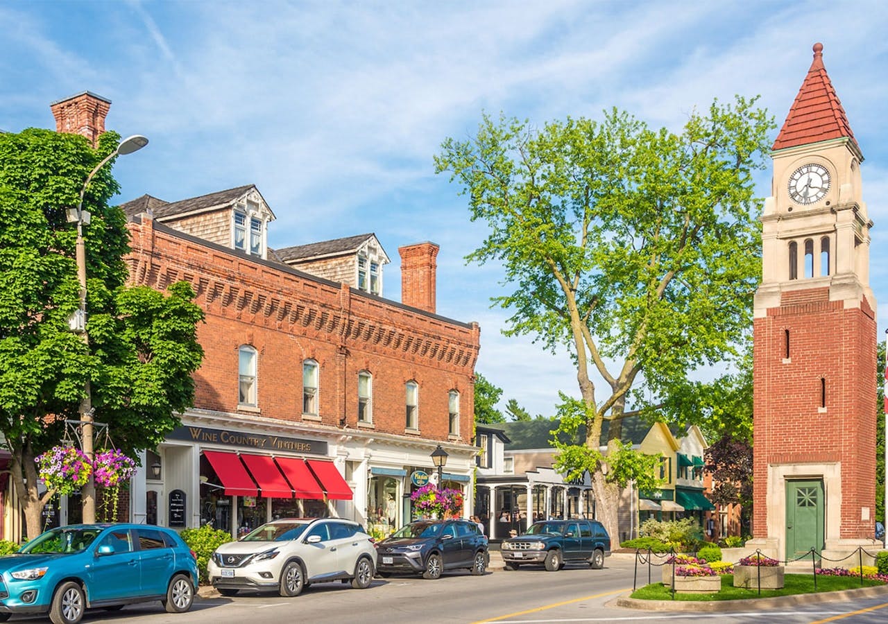 Scène de rue avec des bâtiments en briques, des devantures de magasins, des voitures garées, une grande tour d'horloge, des arbres et un ciel bleu.