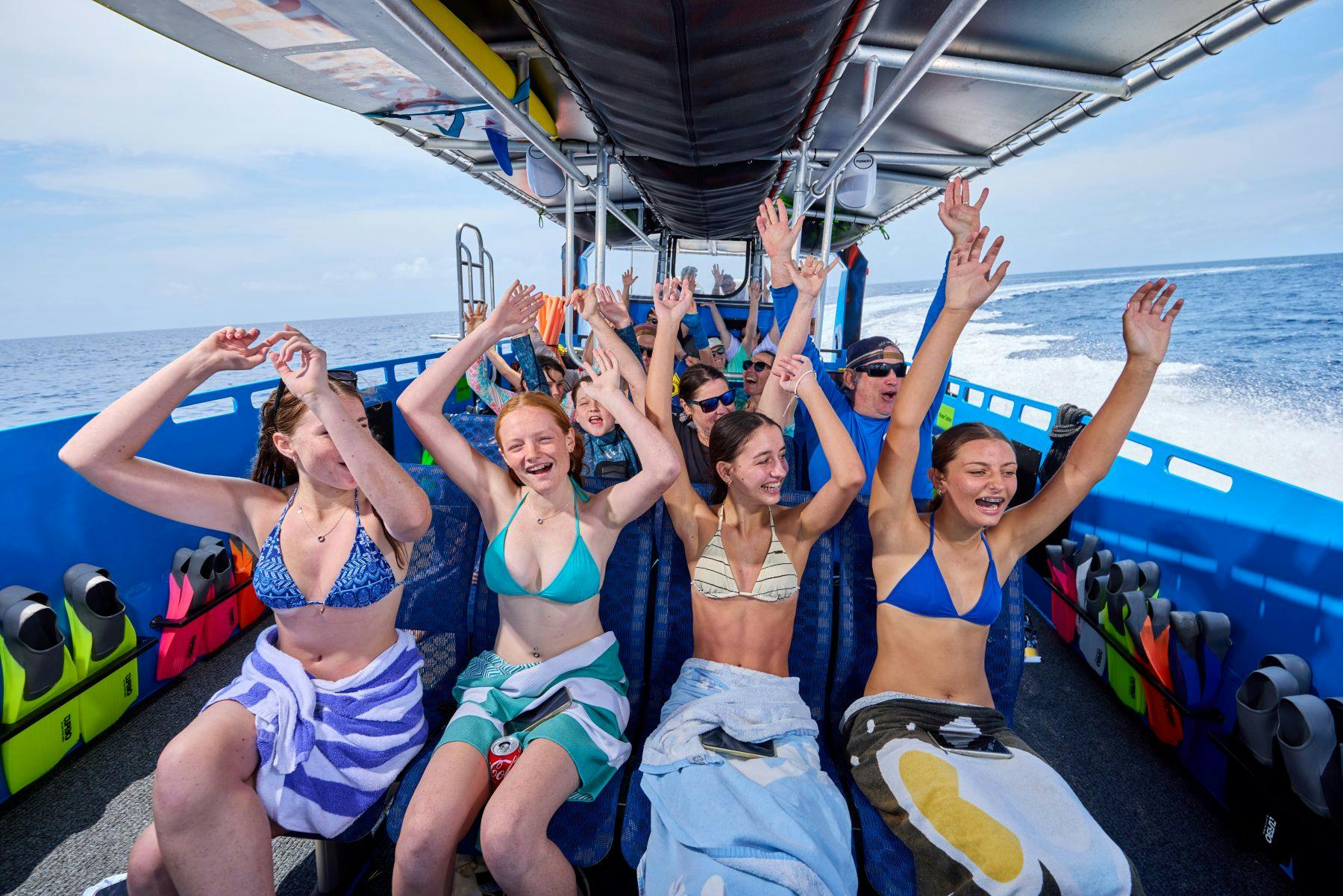 水着とタオルを着た人々のグループがボートに乗っていて、水上をスピードを上げながら笑顔で手を挙げています。