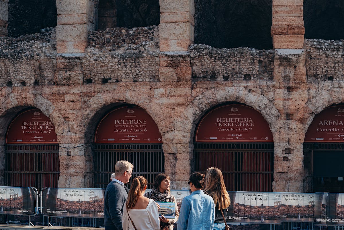 Six people stand and talk in front of an ancient stone structure with ticket office signs.