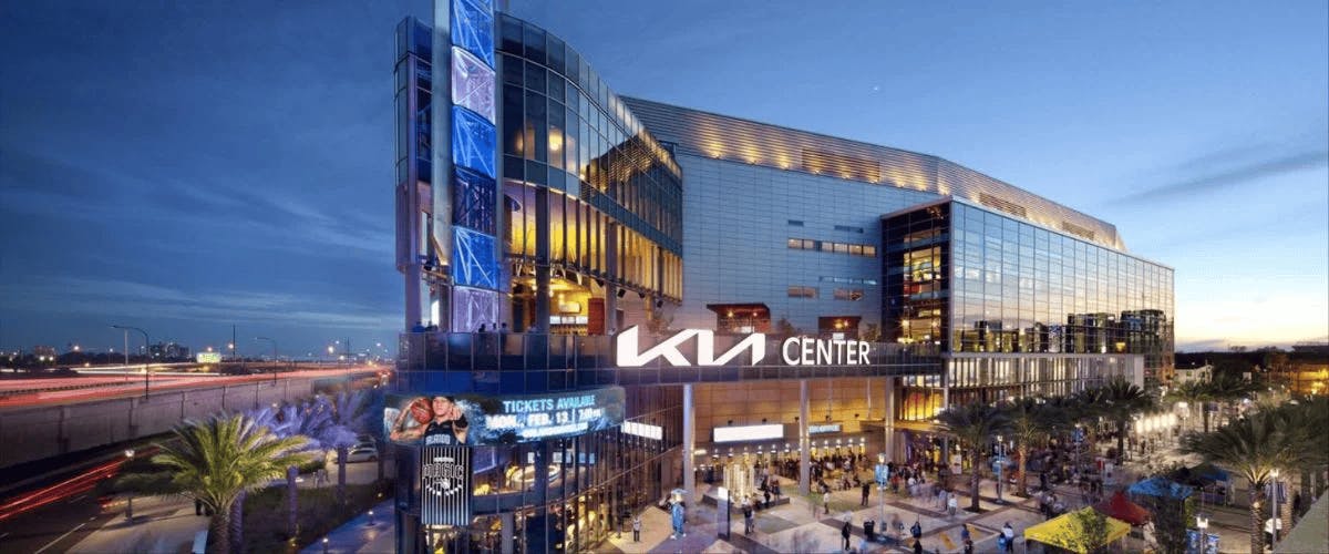 Moderní prosklená budova s nápisem „KIA Center“ za soumraku, s osvětlenými cedulemi, palmami a rušným davem venku.