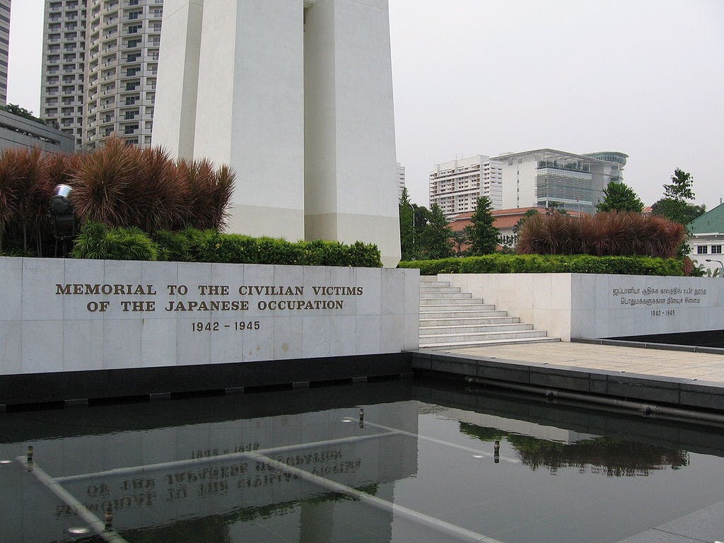 Monument en pierre avec des inscriptions, entouré d'arbustes verts et d'un bassin réfléchissant. L'arrière-plan comprend des bâtiments et des arbres.