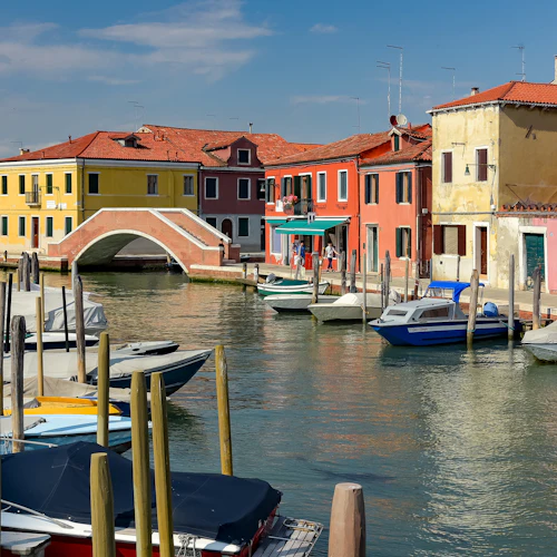 Bunte Häuser am Wasser, Boote, die an einem Kanal angedockt sind, und eine Ziegelsteinbrücke, die beide Seiten unter einem klaren Himmel verbindet.