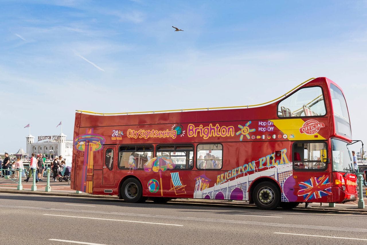 Červený dvoupatrový autobus „City Sightseeing Brighton“ poblíž Brighton Pier s lidmi a ptákem v pozadí pod jasnou oblohou.