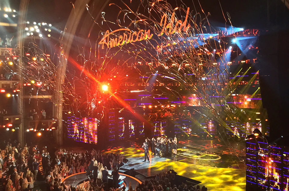 People standing on a brightly lit stage with "American Idol" sign above, surrounded by confetti and an applauding audience.