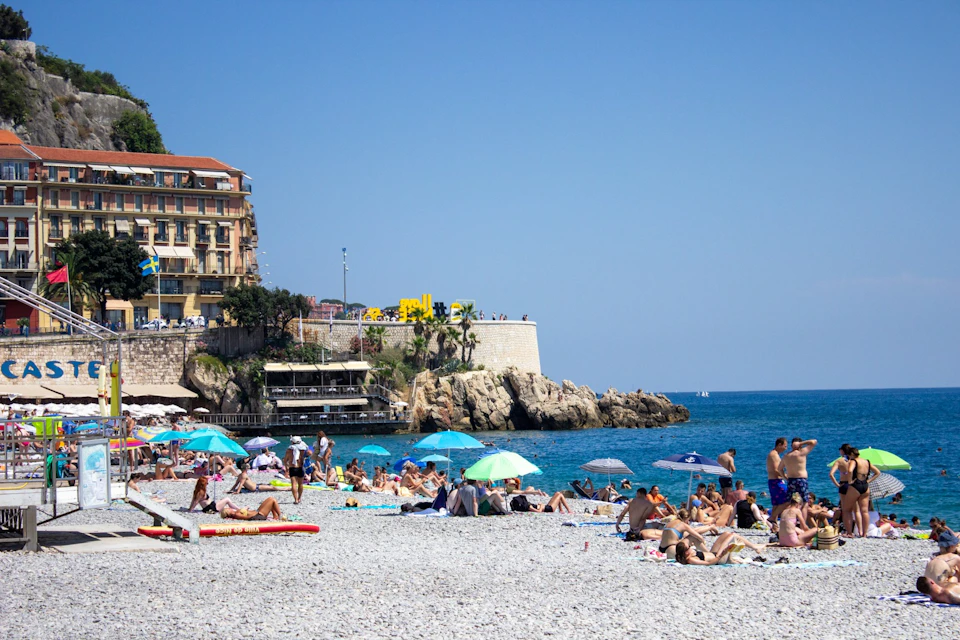 A pebble beach with people relaxing under umbrellas, a rocky shoreline, and a building on a cliff in the background.