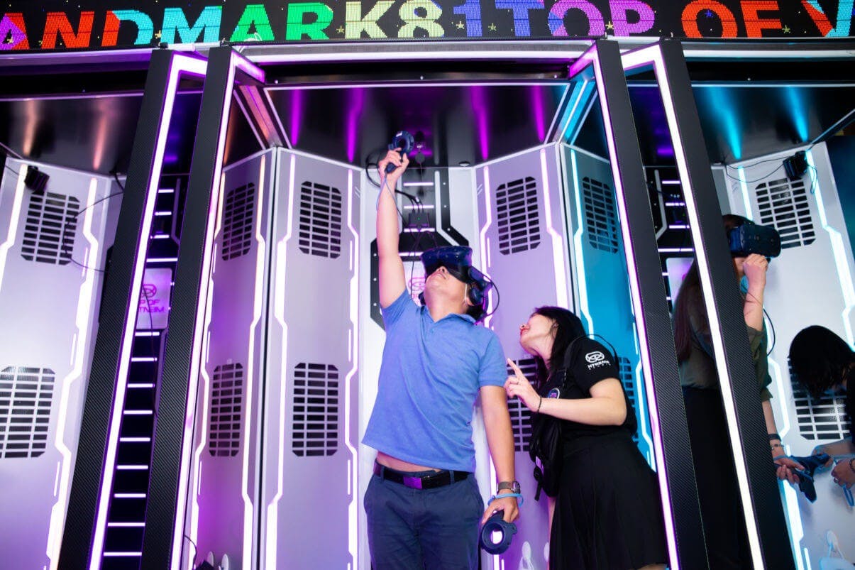A person wearing a VR headset interacts in a futuristic booth, while another stands nearby pointing at the scene. Multicolored text above.