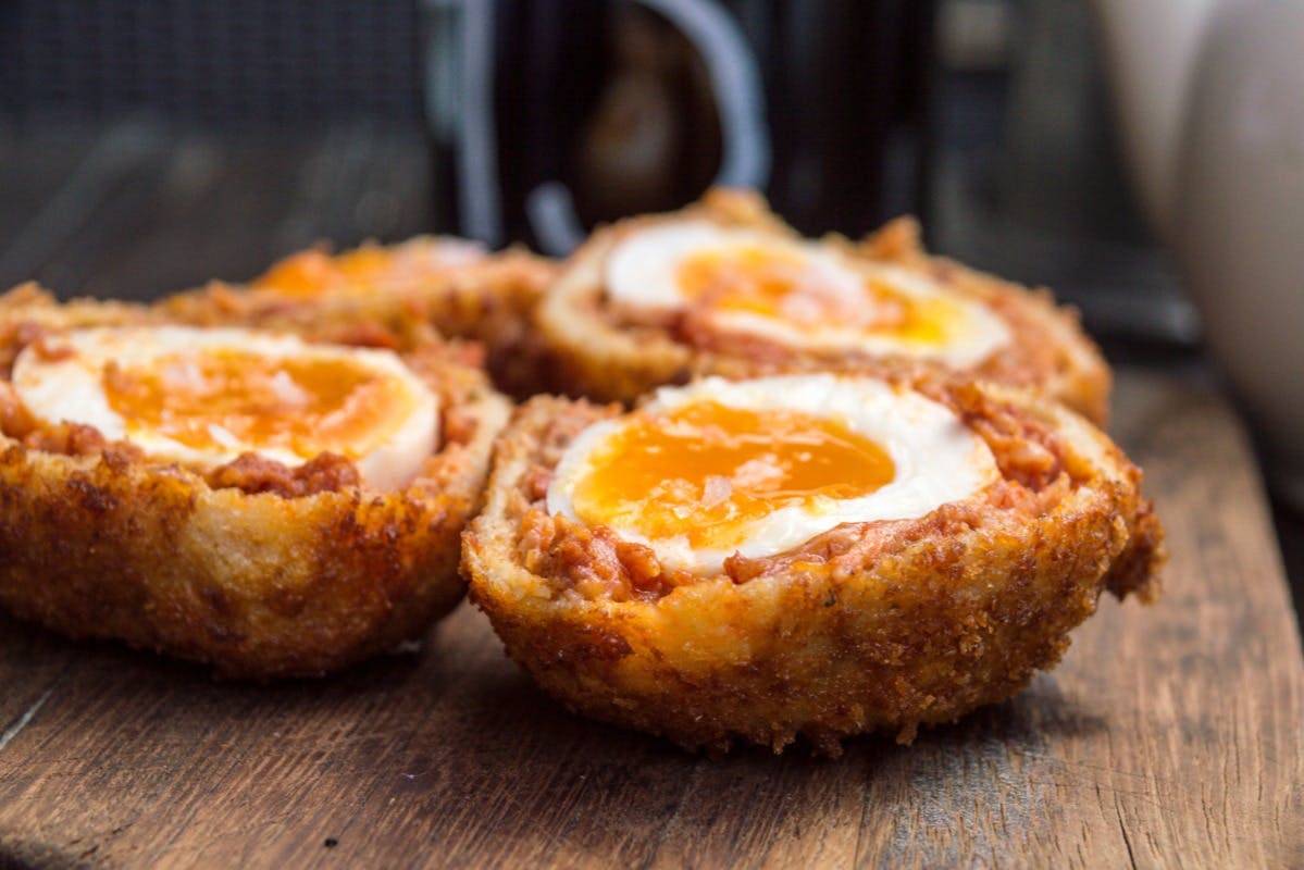 Κοντινό τεσσάρων σκωτσέζικων αυγών σε ξύλινη επιφάνεια, αποκαλύπτοντας μαλακούς κρόκους και τραγανό, χρυσοκάστανο ψωμί.