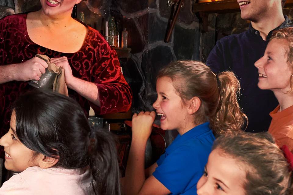 A woman in medieval costume interacts with a smiling man and a group of children in colorful clothing inside a rustic setting.