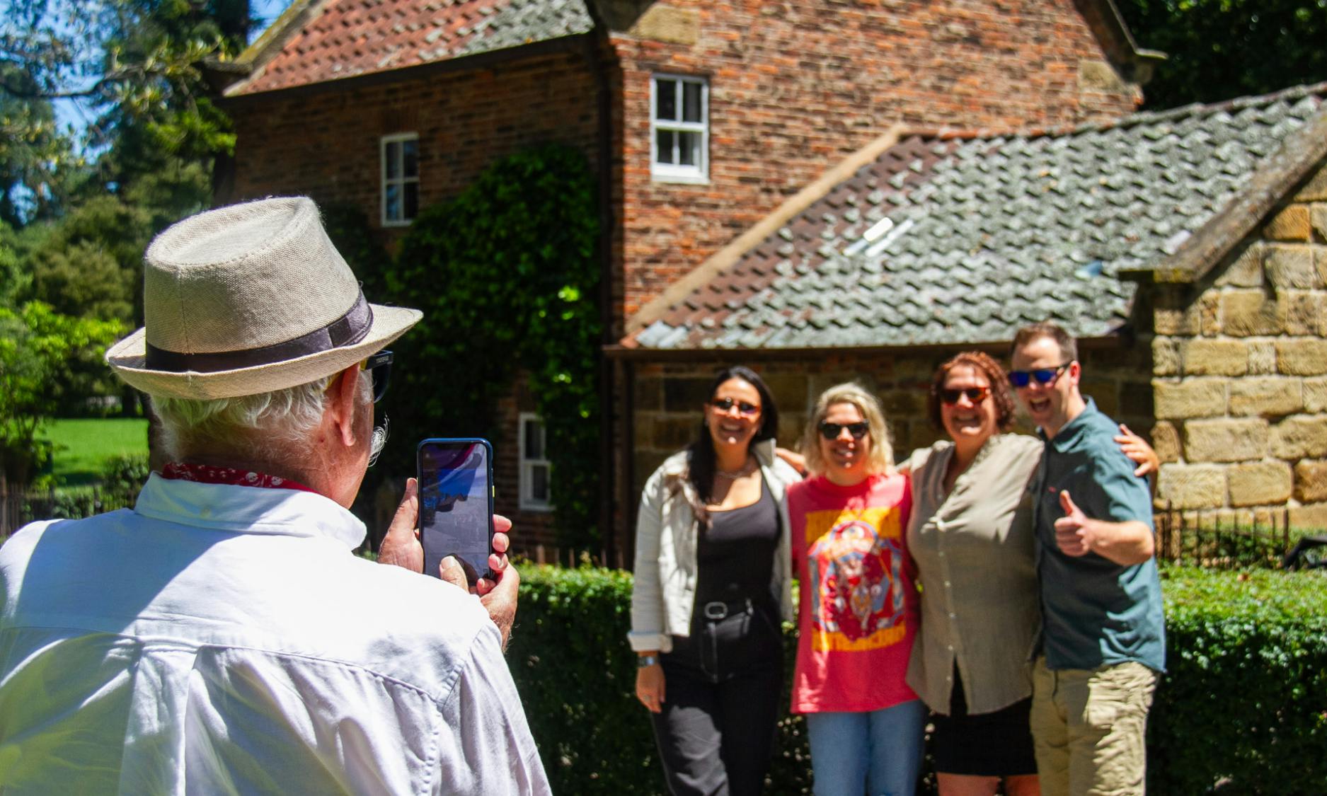 Cztery osoby uśmiechające się i pozujące do zdjęcia na świeżym powietrzu przed ceglanym budynkiem z dachówką. Widoczna jest ręka trzymająca telefon.