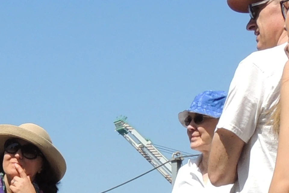 A group of people on a dock, with a uniformed man showing a large, blue torpedo-like object. Cranes are visible in the background.