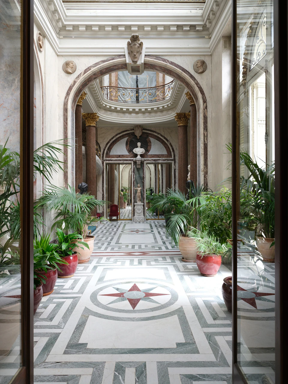 Ornate hallway with marble columns, a statue in the center, and potted plants along the patterned floor.