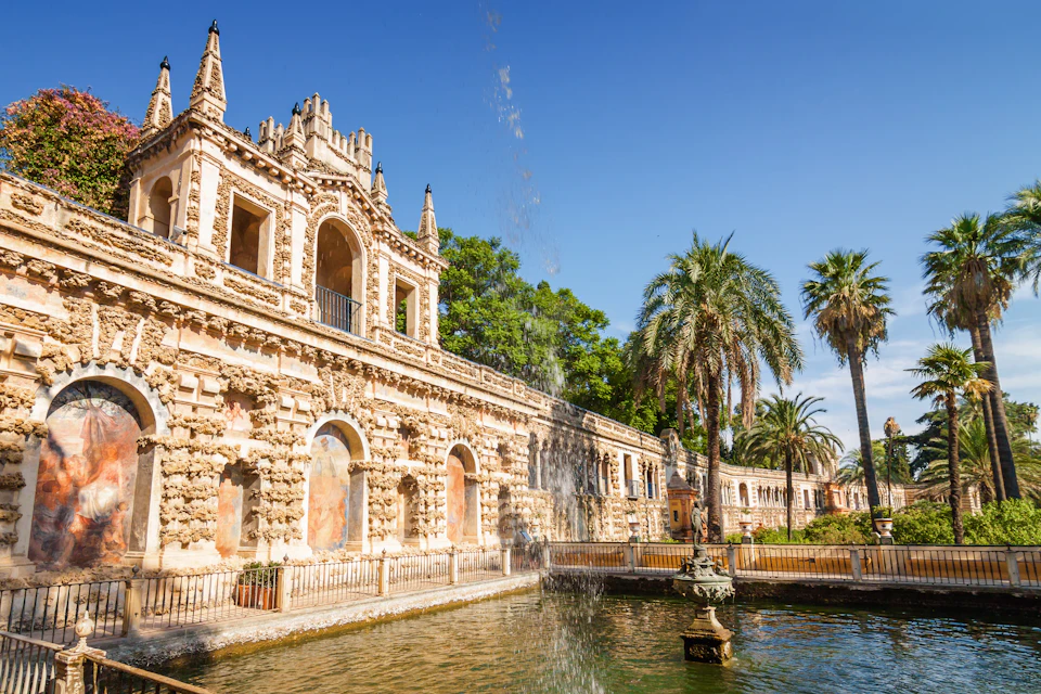 Historic building with intricate architecture behind a water fountain, surrounded by palm trees under a clear blue sky.