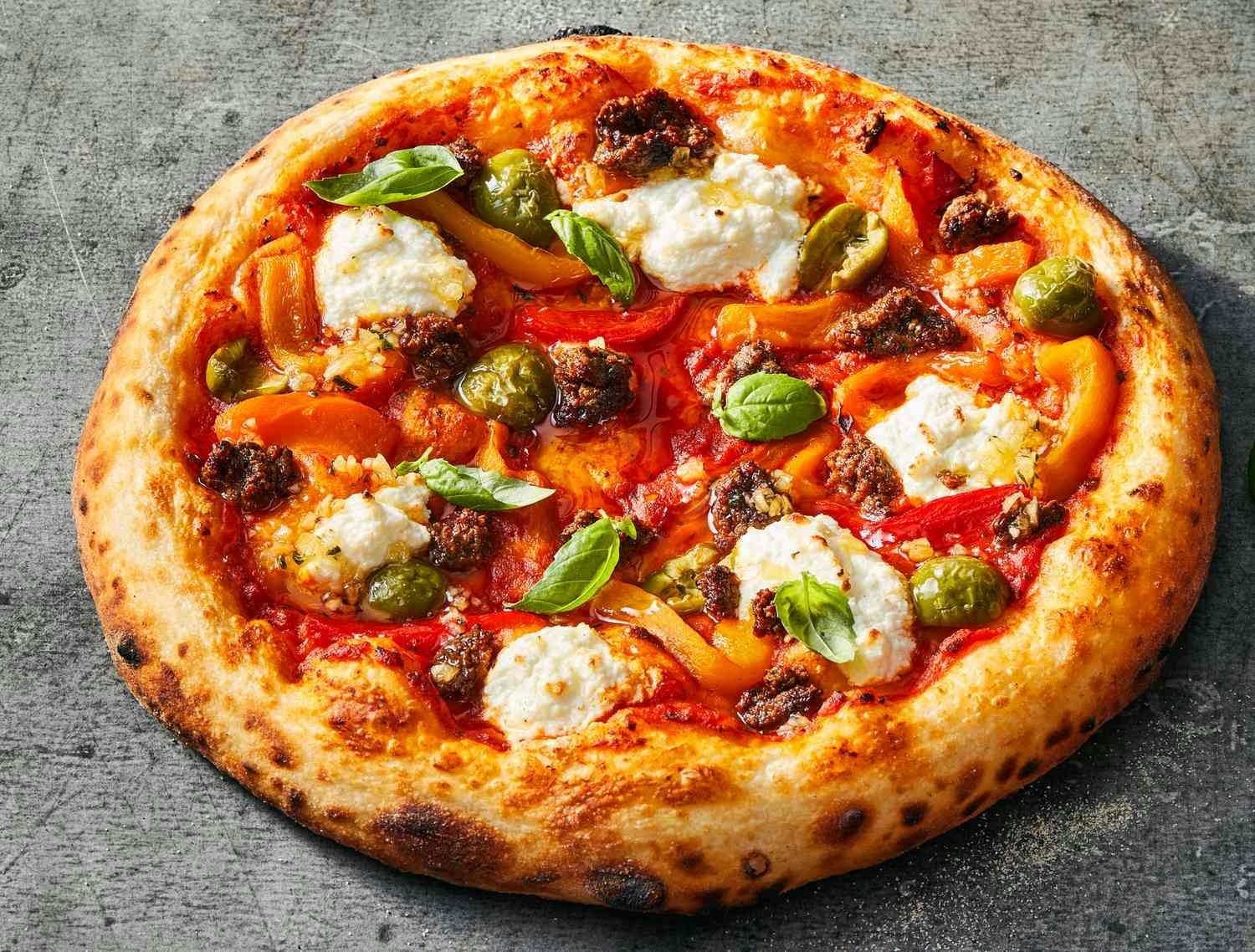 Uma pizza com queijo derretido, azeitonas verdes, folhas de manjericão, carne moída e pimentos vermelhos e amarelos assados numa superfície de pedra.