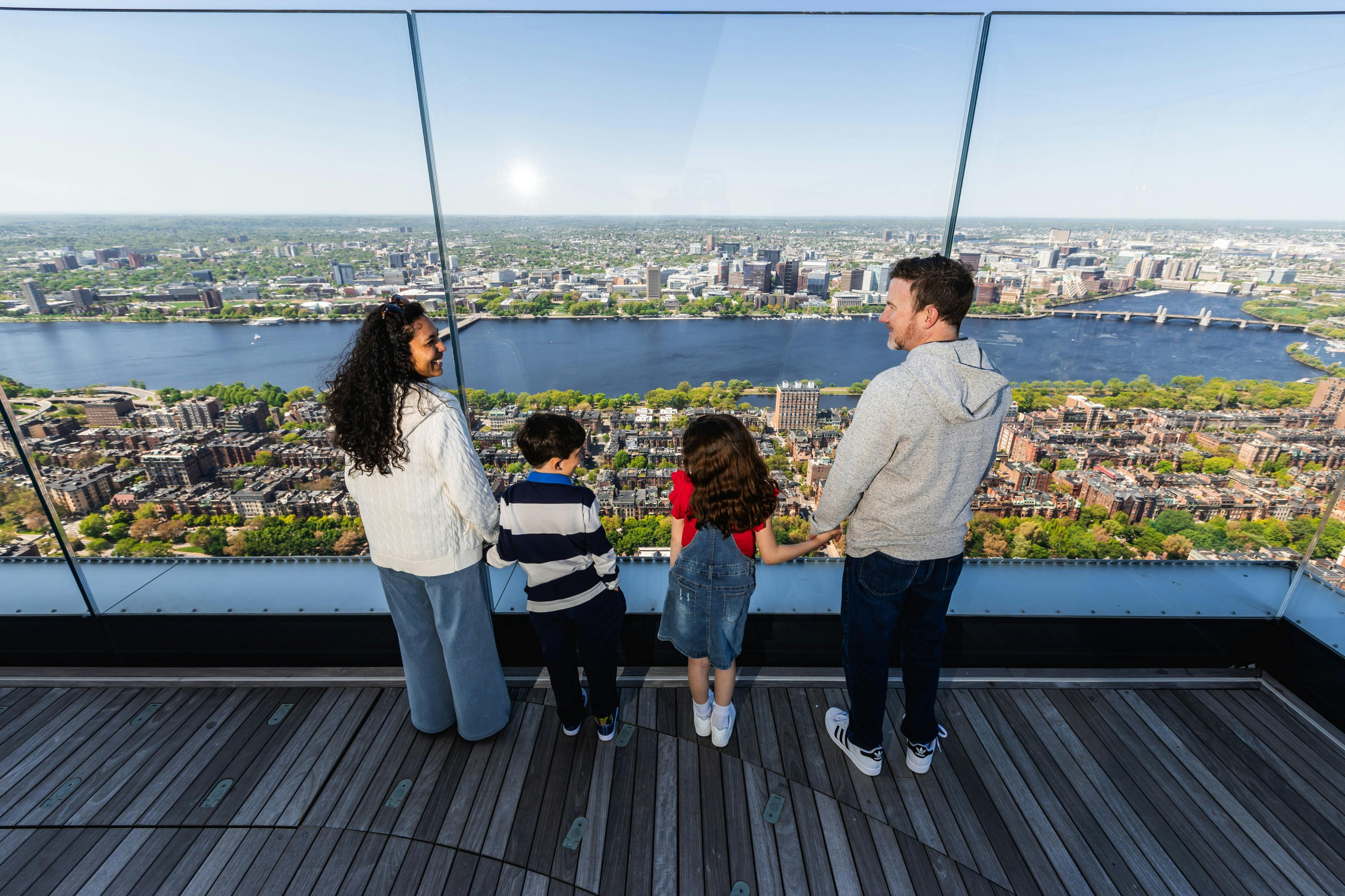 En familj på fyra står på en utsiktsplattform och tittar ut över en stadsbild och en flod under en klar himmel.