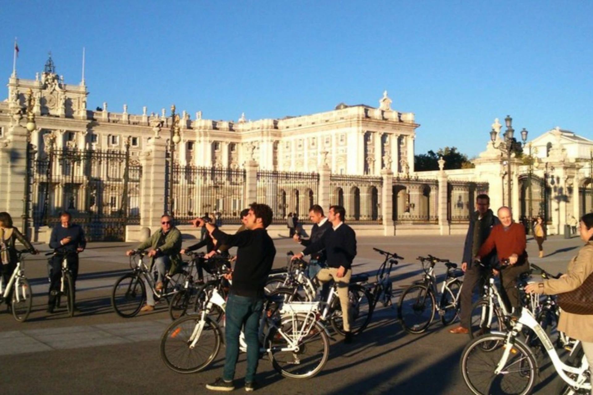 A group of people on bicycles in front of an ornate, white building with a large gate on a clear day.
