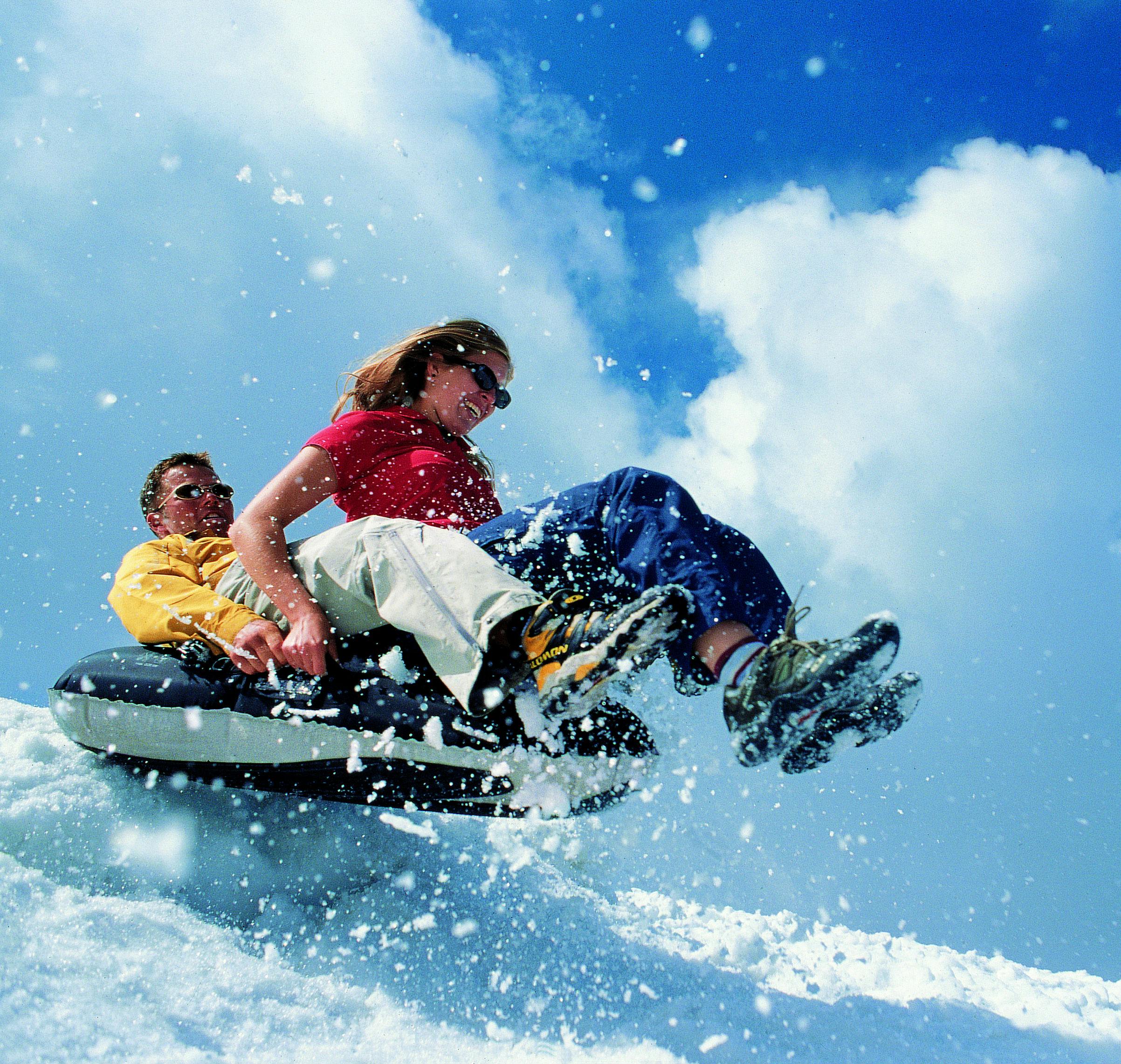 Dva lidé sestupují ze zasněženého svahu, jeden má na sobě červenou košili a druhý žlutou bundu, proti modré obloze.