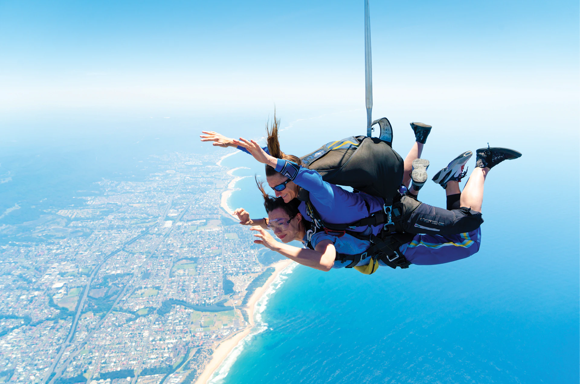 Two people tandem skydiving with a parachute over a coastal city, high above the ocean and shoreline.