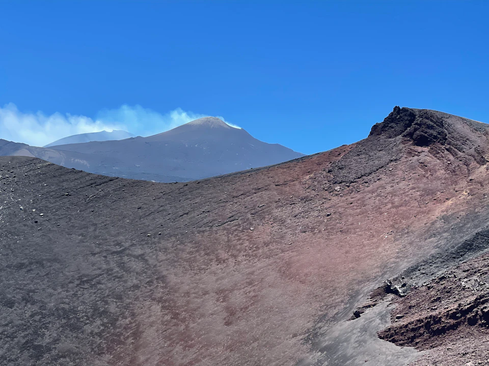 Volcanic landscape with a smoking crater, rugged terrain, and clear blue sky in the background.