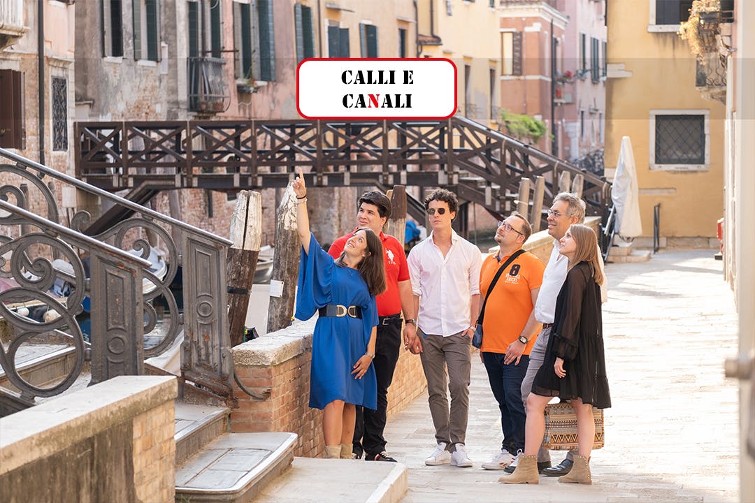 Grup de sis persones caminant per un carrer estret de Venècia, amb una dona apuntant cap amunt. El fons presenta ponts i edificis.
