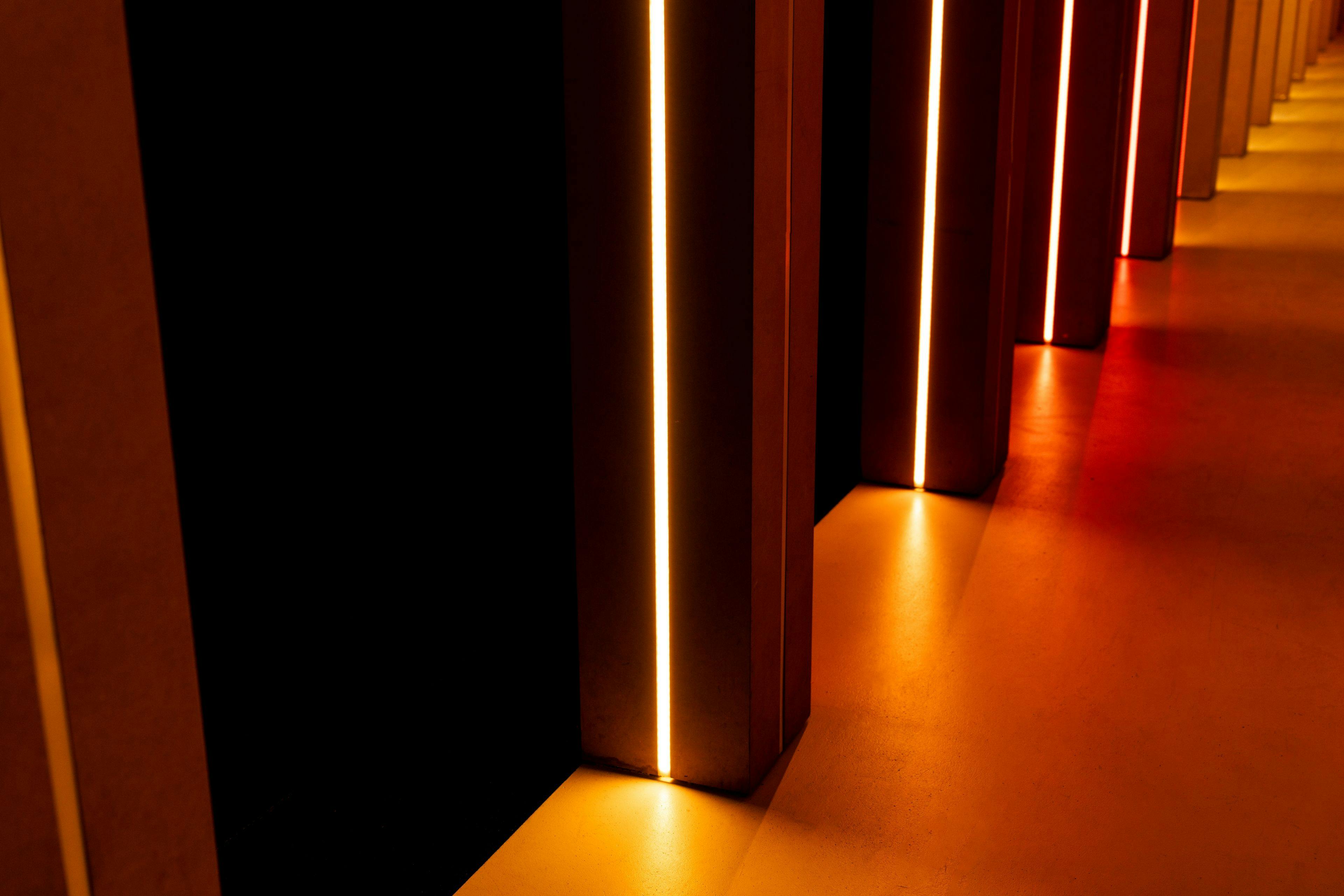Vertikale Balken mit hellen orangefarbenen Lichtern auf dunklem Hintergrund, die warme Reflexe auf einen glatten Boden werfen.