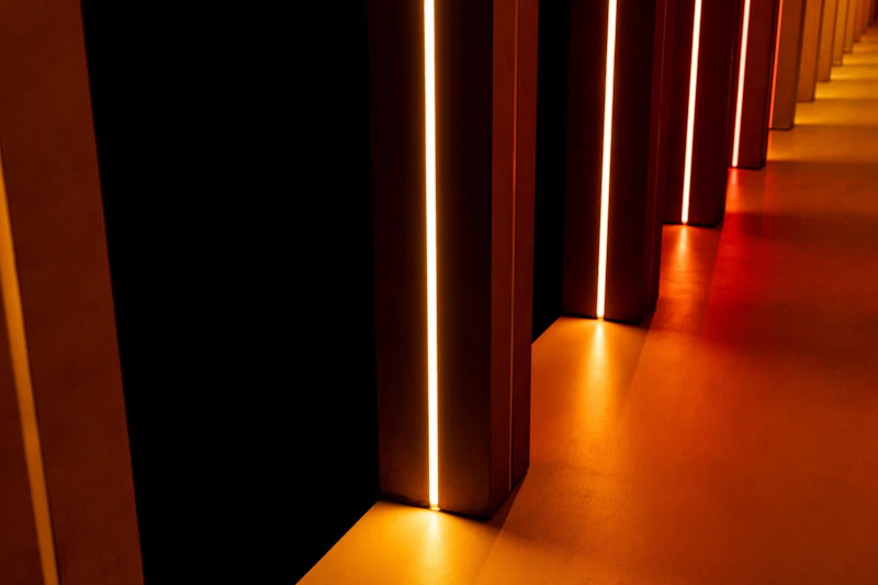Lodrette bjælker med lyse orange lys på en mørk baggrund, der kaster varme refleksioner på et glat gulv.